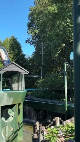 The video shows a water ride attraction at an amusement park. In the foreground, there is a body of water with some greenery and rocks along the edge. Several thick, dark green poles rise vertically, supporting an elevated track. A water ride vehicle, shaped like a boat with a red exterior and a dark interior, travels along the track. The vehicle is carrying two people wearing dark clothing. As the vehicle moves, it creates a significant splash of water. In the background, there are numerous lush green trees and a portion of a building with a dark roof and green siding. The sky is clear and blue. The ride appears to be in motion, as evidenced by the spray of water and the vehicle's movement.