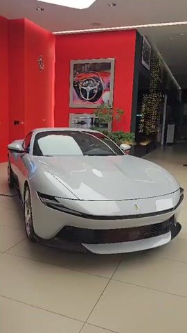 A silver Ferrari is parked in a showroom. The car is the main focus, centered in the frame. The background is a red wall with artwork and framed pictures. To the right, there are more displays and possibly other cars, with decorative lights visible. The floor is tiled. The lighting suggests an indoor, well-lit space.