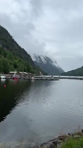 The video shows a serene landscape in Dalen, Norway, featuring a body of water, likely a fjord or lake, framed by majestic mountains. The sky is overcast, with clouds and mist partially obscuring the upper parts of the mountains, creating a dramatic and atmospheric scene. The mountains are covered with lush green trees, with some bare rock faces visible through the vegetation.

In the foreground, a dock extends into the water, with several boats moored alongside. These include various motorboats and smaller vessels like dinghies, gently swaying on the rippled surface of the water. Along the shoreline, multiple wooden buildings, possibly cabins or boathouses, are nestled among the trees. Further down the shore, a larger, more industrial-looking building with a white crane is visible, set against the steep mountainside. The water reflects the cloudy sky and the surrounding landscape, adding depth to the scene. The overall impression is one of peaceful natural beauty amidst a cloudy, cool day.