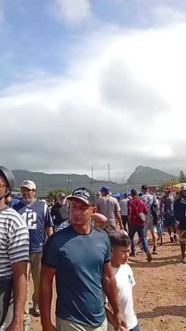 A group of people are walking through an outdoor market under a bright, cloudy sky. The foreground is dominated by a man in a black and white striped shirt, wearing sunglasses and a black crossbody bag. Behind him, other people are visible, including a man in a blue and white jersey and a man in a black baseball cap. In the background, there are stalls displaying produce, including watermelons and pumpkins. Some vehicles are parked in the distance, and a hilly landscape is visible against the sky. The overall impression is a busy, open-air market scene.