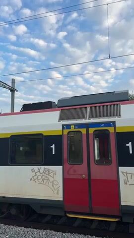 The video shows the side of a white and red train with yellow stripes and black numbering that reads "1". The train is stationary on a track covered with grey gravel. There are red double doors on the side of the train, which appear to be passenger entrances. Graffiti is visible on the lower white portion of the train. Above the train, power lines are visible against a cloudy sky. Trees and a building are in the background. The train is labeled "SBB CFF FFS", which are the initials for the Swiss Federal Railways in German, French, and Italian. The video captures a static view of the train.