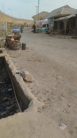 The scene appears to be in a somewhat impoverished area, possibly a rural or developing region. The ground is predominantly dirt and dust, with scattered debris like trash and small rocks. There's an open concrete drainage ditch to the right, filled with visible trash. A small structure with corrugated metal roofing is on the left, possibly a small business or shop, with signage visible. A person stands near the structure. In the background, a wall or fence runs along the left side of the frame, possibly defining the edge of the area. Several vehicles are visible: a car, a motorcycle carrying two passengers, and a cart with a water tank.