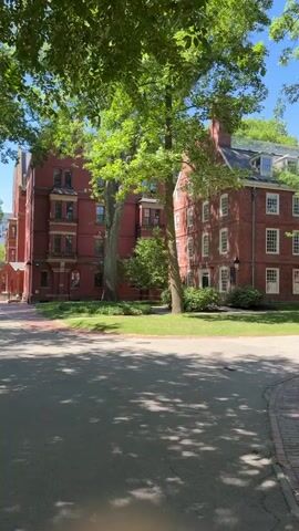 The video begins with a view of a red brick building on a sunny day, surrounded by lush green trees. A brick pathway curves through the foreground, with shadows from the trees dappling the ground. The camera then pans to the right, revealing more of the red brick building, which appears to be a dormitory or academic building, with multiple stories and numerous windows. As the camera continues to move, it captures a wider view of the campus, showing more of the red brick buildings and trees, as well as a few people walking and sitting on the grass. The scene is peaceful and idyllic, conveying a sense of a beautiful summer day on a university campus. One person is seen sitting on a chair under a tree, reading. Another shot shows two people walking past some trash cans. The video concludes with a final panoramic view of the campus, highlighting the architecture and natural beauty of the environment. The overall impression is one of a serene and scholarly atmosphere.