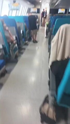 The video shows the interior of a passenger vehicle, possibly a train or ferry, with rows of blue and teal seats on either side of a central aisle. Several passengers are visible. In the foreground, a person in a white top is blurry and walking away from the camera. Further down the aisle, a person in a pink shirt and shorts walks towards the camera, followed by a person in a black shirt. To the left, a young woman with brown hair is seated and looking down at her hands. Other passengers are seated and standing throughout the cabin. Overhead screens display images. The floor is a light gray color.