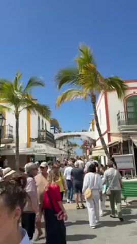 The video shows a crowded street lined with white buildings adorned with orange and red trim under a clear blue sky. Palm trees dot the scene. Many people are walking along the street in both directions. One building has a sign reading "Billy's Apartments."