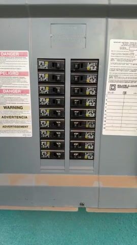 The video shows a close-up of a residential electrical breaker panel. The panel is gray and appears to be mounted on a wall. It contains two columns of circuit breakers, with ten breakers visible in each column. The breakers are black with yellow labels and have levers for turning them on and off. Some of the breakers are in the "on" position, while others are in the "off" position. There are also several warning labels on the panel, including "DANGER" and "WARNING". The bottom of the panel appears dusty or dirty.
