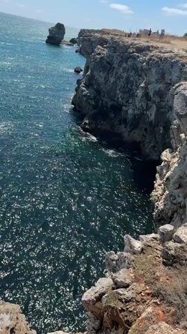 The video shows a rocky coastline with clear blue water. The camera pans across the scene, showing cliffs, rock formations in the water, and the ocean stretching out to the horizon under a bright blue sky. There are a few people visible on the cliff top in the distance. The water is calm with small waves breaking against the rocks. The sun is shining, creating reflections on the water.