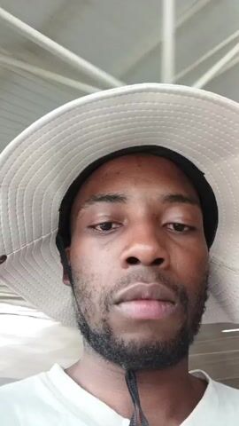 The video shows a close-up shot of a man wearing a white bucket hat. He has dark skin, a beard, and is looking directly at the camera. The background appears to be an airport terminal, with a white ceiling structure visible.