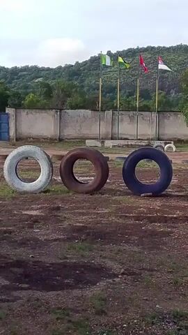 The video shows an outdoor scene with three large tires in the foreground. From left to right, the tires are white, reddish-brown, and blue. Behind the tires is a low, concrete wall. Beyond the wall, there are four flags on poles, each blowing in the wind. From left to right, the flags appear to be Nigeria, Niger, and two other flags that are difficult to identify definitively. In the background, a green, tree-covered hill rises under a cloudy sky. The ground is covered in dirt and some sparse vegetation. The overall impression is of an open, possibly training or recreational area.