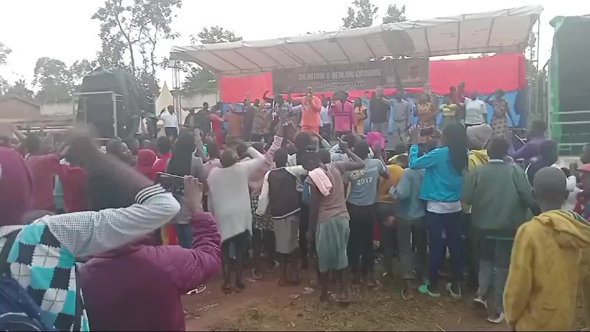 This video captures an outdoor "Salvation & Healing Crusade" event taking place in Matete Market, Kabaruni Nyotei, Lwandeti, Kenya, between October 16th and 18th, 2023.

The scene shows a large gathering of people, predominantly youth and children, engaged in an energetic worship or performance. They are focused on a temporary stage set up with a white canopy roof and red and blue backdrops. A banner prominently displayed on the stage reads "MATETE FOR JESUS SALVATION & HEALING CRUSADE MATETE MARKET 16TH - 18TH OCT 2023 KABARUNI NYOTEI," along with images of individuals, likely event organizers or spiritual leaders.

On the stage, a group of about 15-20 adults, including men and women dressed in various attire, are actively singing and dancing, leading the crowd. Large speakers flank the stage, indicating an amplified sound system. Many in the audience are also dancing, clapping, and raising their hands, demonstrating enthusiasm and participation. Several individuals in the crowd are holding up smartphones, recording the event.

The ground is unpaved, suggesting a market area or open field. Trees and simple structures are visible in the background. The lighting is diffused, indicating an overcast day or possibly late afternoon. The overall atmosphere appears lively and celebratory.