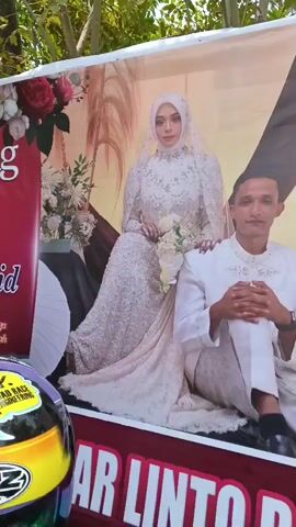 The video shows a large banner of a wedding couple in front of a house. The banner is placed outdoors and is partially obscured by trees and a fence. The wedding couple is depicted in traditional wedding attire. The background of the banner features decorative elements and text. The house in the background appears to be a simple dwelling with a tiled roof and brick walls. A motorcycle is parked in front of the house. The ground is covered with dirt, rocks, and some vegetation. The lighting suggests it is daytime.