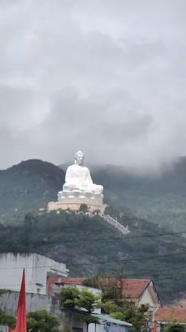 The video shows a large white Buddha statue sitting atop a green hill.  The bottom of the hill is obscured by low-lying clouds.  The foreground shows buildings with red tile roofs and a power pole with many wires.  The day is overcast.