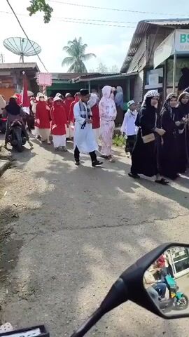 A group of people, mostly children and some adults, are walking in a procession down a paved street. Many of the participants are wearing red outer garments over white undergarments and head coverings, suggesting a uniform or ceremonial attire. Some individuals are holding flags, including one prominent red and white flag which appears to be the Indonesian flag. There are also some people walking in white or light-colored clothing, including one individual in a white robe and headscarf in the foreground. Buildings line the sides of the street, and in the background, a satellite dish is visible. The scene appears to be outdoors, under daylight conditions, with some shadows indicating the sun's position. In the foreground, a motorcycle's side mirror reflects people riding on a motorcycle, with a child sitting between two adults. The overall impression is of a parade or a community event.