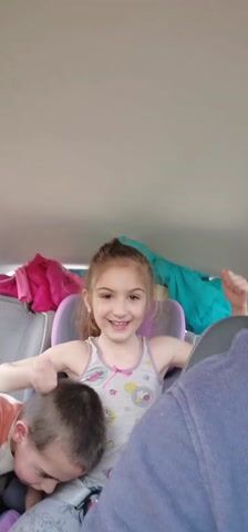 The video shows a girl sitting in a car, giving a thumbs-up and smiling. She is wearing a tank top and has her hair in a ponytail. There are two other children in the back seat of the car. The boy on the left is leaning forward with his head down, and the girl on the right is leaning against the car seat. There are coats and other items piled up behind them in the back of the vehicle. A man, the recorder of the video, is in the front of the car. The interior of the car is visible, and music is playing in the background.