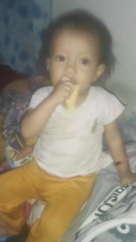 A baby is lying down in bed. The baby is wearing a white t-shirt and orange pants. The baby is holding a yellow object and putting it in their mouth. The baby is looking at the camera. The baby’s head is tilted back, and their mouth is open. The baby’s eyes are wide open, and they are looking at the camera. The baby’s body is relaxed, and they appear to be comfortable. The room is dimly lit, and the walls are white. There is a blanket on the bed with a pattern on it.