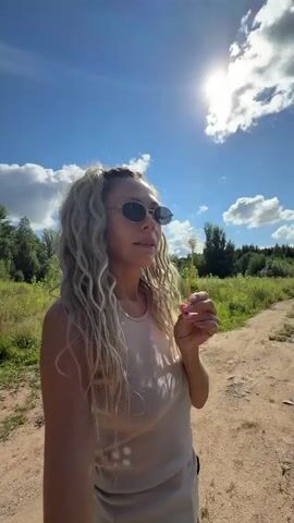 A woman with blonde curly hair and sunglasses is outside blowing on a dandelion. The sun is shining brightly in a clear blue sky with some white clouds. The woman is wearing a light-colored tank top and is holding the dandelion stem with her right hand. She is standing on a dirt path with green foliage and trees in the background.