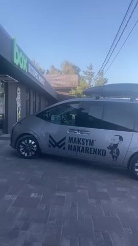 A silver minivan with the words MAKSYM MAKARENKO DENTAL CLINIC and a cartoon image of a man on the side is parked in front of a building. The building has a green sign that says BOX. The minivan has a roof rack with a cargo box. The ground is paved with dark bricks. The sky is clear and blue. The time is early morning or late afternoon, as indicated by the lighting.