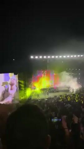 The video shows a live concert at night. The main performer is on stage, singing into a microphone. He's wearing a dark shirt and jeans. There are other musicians on stage, including a drummer behind him and a man to his left holding a camera. 

Behind the musicians is a large screen displaying colorful images of the performer's face in a pop-art style. There are bright stage lights illuminating the scene, and some smoke or fog in the air.

In the foreground is a large crowd of people, mostly visible from the chest up. Many are holding up their phones to record the concert. The video is taken from within the crowd, slightly obstructed by a head.