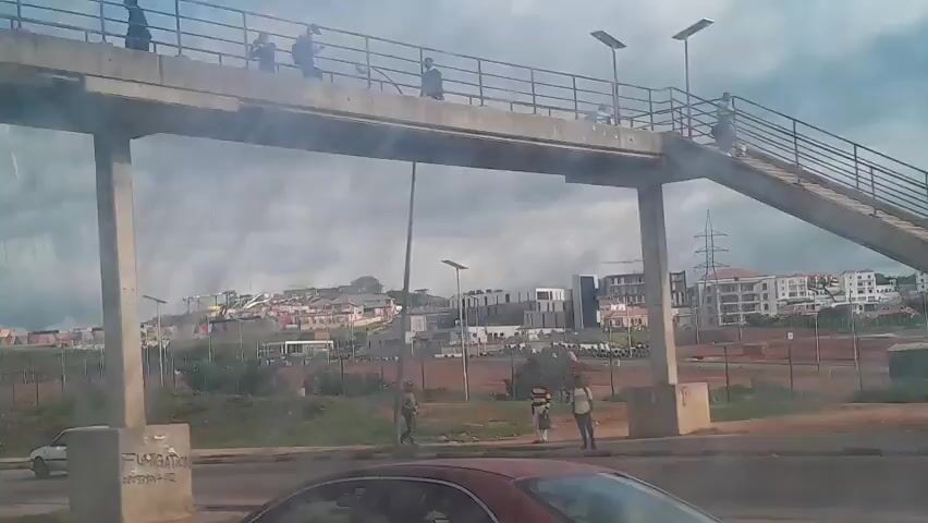 The video shows an outdoor scene with a road in the foreground and buildings on a hill in the background. A pedestrian bridge passes overhead in the first few seconds. Several cars are visible on the road, including a white hatchback and part of a red car. There are a few pedestrians on the ground and on the pedestrian bridge. The sky is cloudy. The scene appears to be a developing urban area with construction activity visible.
