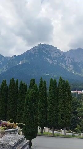 The video begins with a view of a majestic mountain range under a cloudy sky. In the foreground, a line of tall, slender cypress trees stands against a backdrop of lush green foliage and distant buildings. As the camera pans slightly, the scene transitions to reveal more of the surrounding park-like environment. There is a paved pathway with a lamppost and manicured bushes. Further into the frame, two individuals are visible standing near a decorative white frame, possibly posing for a photograph. The overall impression is a serene and picturesque landscape, likely a tourist destination or a well-maintained park, with the imposing mountain range serving as a dramatic natural element.
