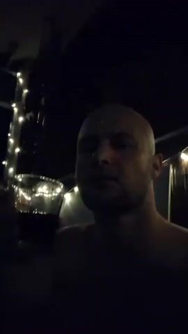 A man with a shaved head is holding a glass of red wine to his lips. He is partially submerged in dark water, and the scene is lit by string lights. The man appears to be in a pool or hot tub.