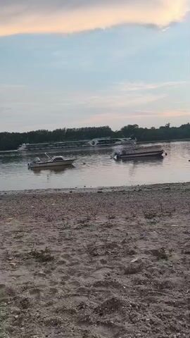 The video shows a serene body of water with several boats anchored near the shore. The sky above is a soft blend of blue and pastel hues, suggesting either sunrise or sunset. The foreground consists of a sandy beach with scattered pebbles and sparse vegetation. The overall ambiance is tranquil and peaceful.