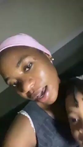 A woman with a light pink headband, wearing a grey tank top with white lace trim, holds a toddler in her arms. The toddler wears a sleeveless striped top and is looking at the camera. The woman is singing along to music and appears to be recording a video. The background is slightly blurred, but shows dark walls and some objects like a blue container.