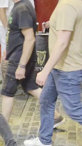 A man in a tan t-shirt and blue jeans walks past a Guinness barrel being used as a table. Three other men stand around the barrel. One man on the left wears a white t-shirt and light colored shorts. The second man from the left wears a white t-shirt and black shorts. The man on the far right wears a white t-shirt and denim shorts. Two glasses of dark beer and a can of what appears to be soda sit on the barrel. The ground is paved with cobblestones.