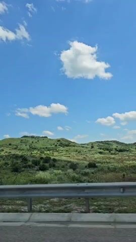 A car passenger's point of view, looking out of the window at a green, rolling landscape under a bright blue sky with scattered white clouds. A metal guardrail is visible in the foreground, and part of the road surface. A blue directional sign appears briefly, indicating directions to "A-157 AIRPORT" and "GENERALNIE VODY," as well as "LERMONTOV" and "ESSENTUKI" with a distance of 400m. The sign also has directional arrows and what appear to be circular symbols. The landscape is characterized by lush green vegetation, with some trees and bushes dotting the hills. The time of day appears to be daytime, likely sunny.