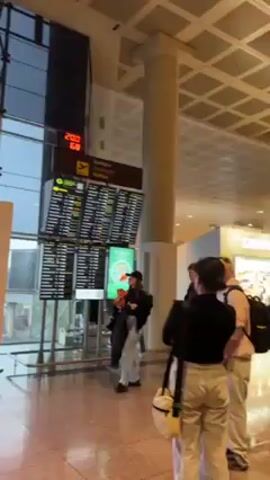 The video shows a group of people standing in front of a departures board at an airport. The board is displaying flight information, including destinations, flight numbers, gate numbers, and status updates. The people are looking at the board, presumably checking for information about their flights. The airport is modern and well-lit, with a high ceiling and large windows. The floor is tiled. The airport is busy, with people walking around and waiting for their flights. The camera zooms in on the departures board, showing the flight information in more detail.