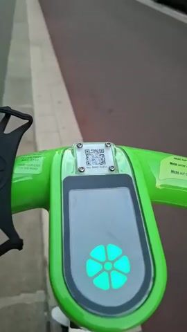 The video begins with a close-up of a green electric scooter's handlebars. A QR code is visible on a small display above a larger screen that shows a green, flower-like symbol. The perspective then shifts to a street-level view, looking down a street lined with buildings and a sidewalk. A red-brick church with a tall steeple is visible at the end of the street. The street appears to be paved with dark asphalt and has a lighter gray border. Cars are parked along the right side of the street. The sky is overcast. The scooter is moving forward, indicated by the changing perspective and the blurred motion of the surroundings. A person is briefly visible on a scooter further down the street.