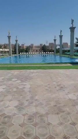 A wide shot of an outdoor swimming pool on a clear, sunny day. The pool is rectangular and filled with blue water. It is surrounded by a green, grassy area, and beyond that, a patterned paved walkway. In the background, there is a long, low building with a crenelated roofline, resembling a castle or fortress. Several tall, white pillars topped with golden statues stand along the sides of the pool. Lounge chairs are visible behind the pool and around the building. The sky is a clear, bright blue.