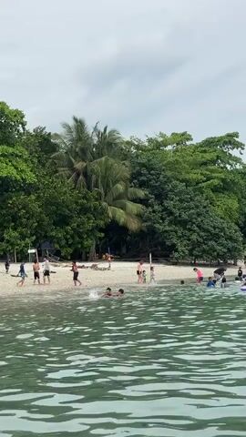 A static medium shot captures a group of people enjoying themselves at a beach. The camera is positioned from the water, looking towards the shore. The water in the foreground is a light green with visible ripples. Several children are in the water, some wearing inflatable rings, while others are playing closer to the shore. On the sandy beach, adults and children are walking, standing, and interacting. The beach is backed by a dense line of various green trees, including palm trees. The sky above is overcast with a mix of grey and white clouds. The overall scene suggests a leisurely day at a tropical beach.