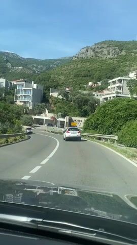 The video captures a view from a vehicle traveling on a paved road that winds through a hilly, green landscape. Several buildings, appearing to be hotels or residences with modern architecture, are visible on the hillsides. The road has lane markings and guardrails. Other vehicles, including a white Fiat, a dark sedan, and other cars, are also on the road, some ahead and some approaching. The sky is blue with scattered white clouds, suggesting it is daytime and fair weather. The overall impression is of a scenic drive through a coastal or mountainous tourist area.