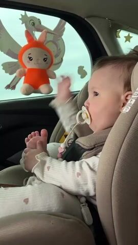 A baby girl with a pacifier in her mouth sits in a car seat. She is wearing a beige patterned onesie with frills on the sleeves. She is holding her left foot in her hands and playing with it. There is an orange bunny stuffed animal hanging from the car window with a stork. The car seat is beige and has an airbag warning tag. The car is in motion and the background is blurred.
