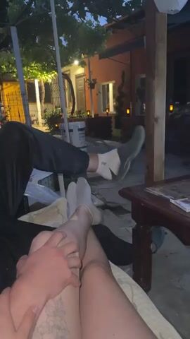 A close-up shot of a person's legs and hands, with the focus on two people's legs intertwined and a hand resting on a leg. The setting appears to be an outdoor patio or garden area, possibly at dusk or in the evening, as indicated by the dim lighting and visible lamps in the background. One person's legs are extended, with one foot resting on a wooden surface, while the other person's legs are bent, with a hand placed gently on their thigh. The background shows a building with warm lighting, trees, and vegetation, suggesting a relaxed, intimate atmosphere.