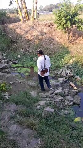 A person wearing a white long-sleeved shirt, dark pants, and a head covering walks along a rocky, overgrown creek bed. A brown bag is slung across their back. The environment appears to be rural, with trees and vegetation visible in the background. The creek water is murky and has patches of green algae.