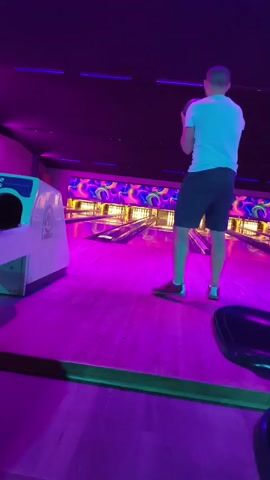 A young adult male wearing a white t-shirt and black shorts stands on a purple-lit bowling lane, holding a dark bowling ball. He moves to bowl, swinging his arm back and releasing the ball down the lane. The lane is lined with pins and has a neon-colored design on the back wall. A white bowling ball return machine is visible on the left side of the frame.