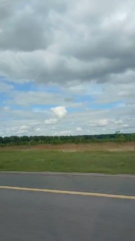 The video shows a landscape with a cloudy sky and a field of green grass. A road runs in the foreground with a yellow line marking the lane. In the distance, there is a line of trees. Several contrails are visible in the sky, suggesting that airplanes have recently flown overhead. The weather appears to be partly cloudy with some sunshine breaking through. The scene is serene and peaceful, with no people visible. The video captures a fleeting moment of natural beauty as the viewer moves along the road.