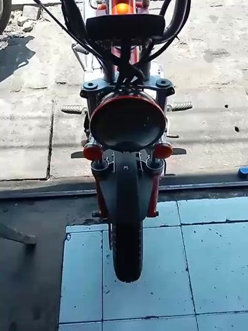 The video shows a close-up view of a red motorcycle from a slightly elevated angle, focusing on the front and handlebars. The motorcycle is stationary, parked on a tiled surface. The environment appears to be a workshop or garage, with concrete flooring visible in the background and a hint of another motorcycle to the left. The motorcycle itself features a round headlight, orange turn signals, and black handlebars with brake levers. A label reading "SPEEDHUNTERS" is visible on the side of the fuel tank. The overall lighting suggests it is daytime. There are no people or any visible actions taking place in the video.