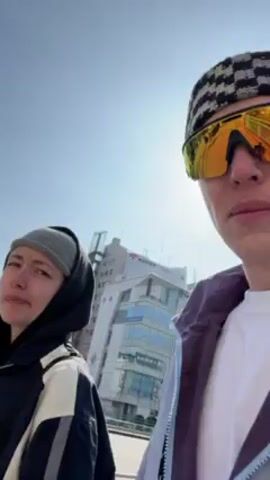 Two young adults stand outdoors on a bright, sunny day. The person on the right is wearing a checkerboard beanie and mirrored sunglasses, and is closer to the camera. The person on the left wears a gray beanie and a black hooded jacket with a white stripe on the sleeve. Behind them are several tall buildings under a clear blue sky.