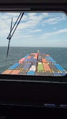 A first-person perspective video shows the view from the bridge of a container ship at sea. The camera pans across the deck of the ship, which is laden with colorful shipping containers, and then out towards the ocean. The sea is choppy, with whitecaps visible under a bright, partly cloudy sky. The video also shows some of the ship's navigation equipment, including screens and control panels. The overall impression is one of a ship sailing through open water.