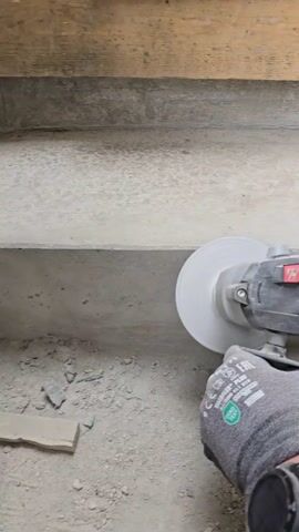 A gloved hand holds a running power tool with a large disc-shaped attachment against a grey concrete surface. The concrete surface appears to be steps. Debris lies on the ground near the steps. The power tool is dark blue and black, with a red on/off switch. The glove is grey and has a green circular label with “OEKO-TEX” printed on it, among other things. The power tool is being used to smooth or grind the edge of a step.