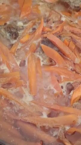 The video shows a close-up view of sliced carrots being cooked in a simmering liquid in a pot. Steam rises from the surface. Darker bits, possibly cooked meat or other vegetables, are visible amongst the carrots. Bubbles form in the liquid.