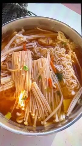 The video opens with a close-up of a bowl of ramen noodles with enoki mushrooms and carrots in a broth. The camera then pans out to show the bowl on a portable gas stove outdoors, with a body of water and grassy area in the background. A person wearing a black t-shirt, black shorts, and a black baseball cap enters the frame, bending over and appearing to be interacting with something on the ground. The scene suggests an outdoor cooking setup, possibly near a beach or lake, given the presence of water and sand-like ground.