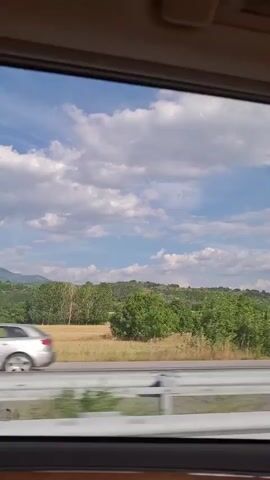 This is a point-of-view video from inside a vehicle, looking out the side window. The landscape outside is visible, featuring a bright blue sky with scattered white clouds. Below the sky, there are rolling hills covered in green trees and patches of dry, golden vegetation. A road is visible in the foreground with a white barrier. At the 00:01 mark, a silver car passes by in the opposite direction on the road. The interior of the vehicle is dark and out of focus, framing the view. The overall impression is one of a scenic drive through a rural area.