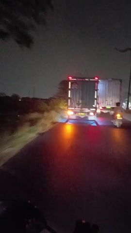 This video captures a nighttime journey along a rural road in Wangunharja, Indonesia. The primary subjects are a motorcyclist, who is the perspective from which the video is filmed, and two large trucks ahead. The scene is dimly lit, suggesting it's late evening or early morning, with the only significant light sources being the headlights and taillights of the vehicles and a distant streetlight.

The motorcyclist is riding on a paved road, with the left side of the road featuring a grassy embankment and sparse vegetation, some of which appears to be trees. The right side of the road is occupied by the two trucks, one in front of the other, both appearing to be container trucks. The truck in front has its taillights illuminated, showing red and white lights, and it's difficult to discern any specific markings. The truck behind it is closer and more clearly visible, with a metallic container. Its taillights are also red, and there's a blue glow emanating from underneath the rear of this truck, possibly from aftermarket lighting. A person, presumably the motorcyclist, is visible in front of the camera, riding a scooter or motorcycle. Their silhouette is visible against the light of the truck's taillights.

The activity is a simple journey or commute on a road. The environment appears to be relatively quiet, with no other vehicles clearly visible apart from the two trucks and the motorcyclist. The sky is dark, with no stars visible, suggesting cloudy weather or heavy light pollution. The overall mood is one of a solitary nighttime ride, with the bright lights of the trucks providing a focal point in the otherwise dark surroundings. There is no discernible text visible in the video.