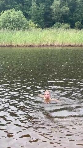A person is swimming in a body of water. They are partially submerged, with their head and shoulders visible above the surface. They appear to be moving their arms and legs as if swimming. The water is dark and choppy, with ripples on the surface. In the background, there is a shoreline lined with tall, green reeds. Behind the reeds, there is a dense forest with trees. The lighting suggests it is daytime.