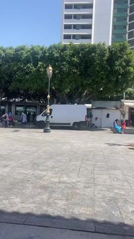 The video shows a public square in a city. The square is paved with gray tiles. There are a few trees with dense green foliage scattered throughout the square. A white van is parked near one of the trees. Several people are sitting on benches or walking around. In the background, there are tall buildings, one of which is white with green horizontal stripes. The sky is clear and blue.