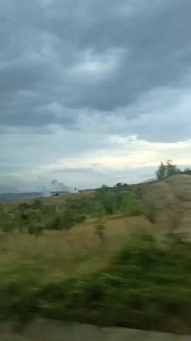 The video shows a landscape from a moving vehicle, likely a car or train, looking out of a window. The scene is dominated by a cloudy sky and rolling hills covered in green and brown vegetation. In the distance, there is a plume of smoke rising, suggesting some kind of industrial activity or a fire. The camera is angled slightly downwards, showing a road or barrier in the foreground. The overall mood is somewhat overcast and potentially somber due to the smoke.